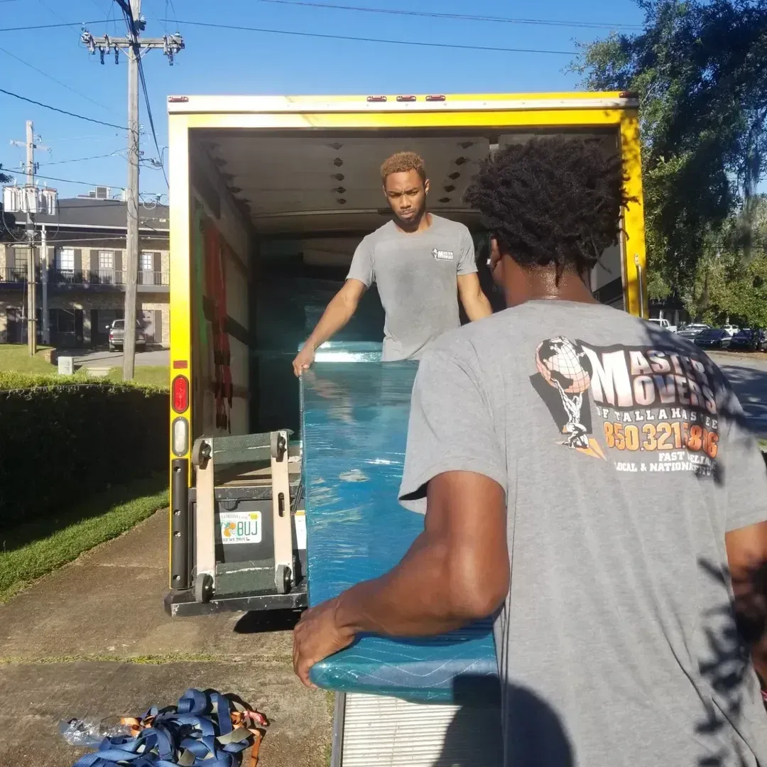 Master Movers of Tallahassee workers unload a blue mattress from a box truck on a sunny street.