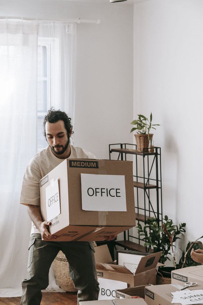 Man carrying a cardboard box labeled "OFFICE" in a room with other boxes, moving.
