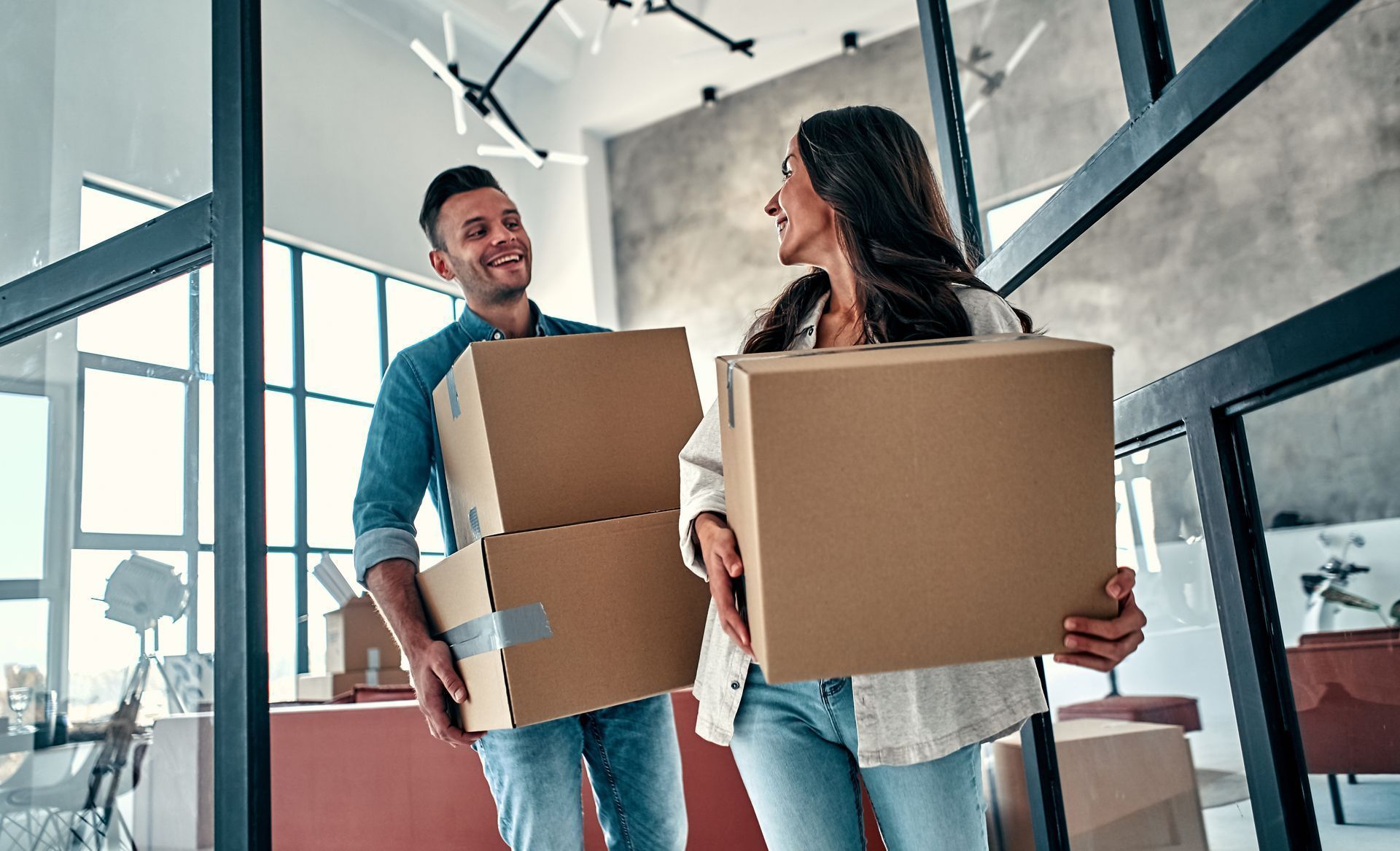 Couple carrying cardboard boxes, smiling, inside a new apartment.