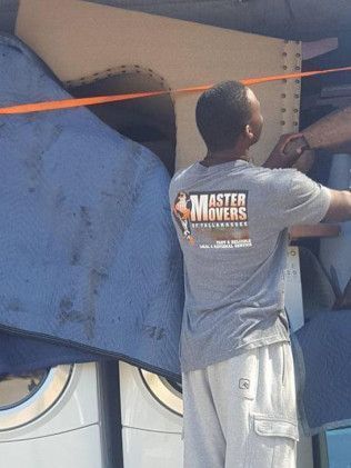 Man in "Master Movers" shirt securing items in a truck, blue padding, and appliances visible.
