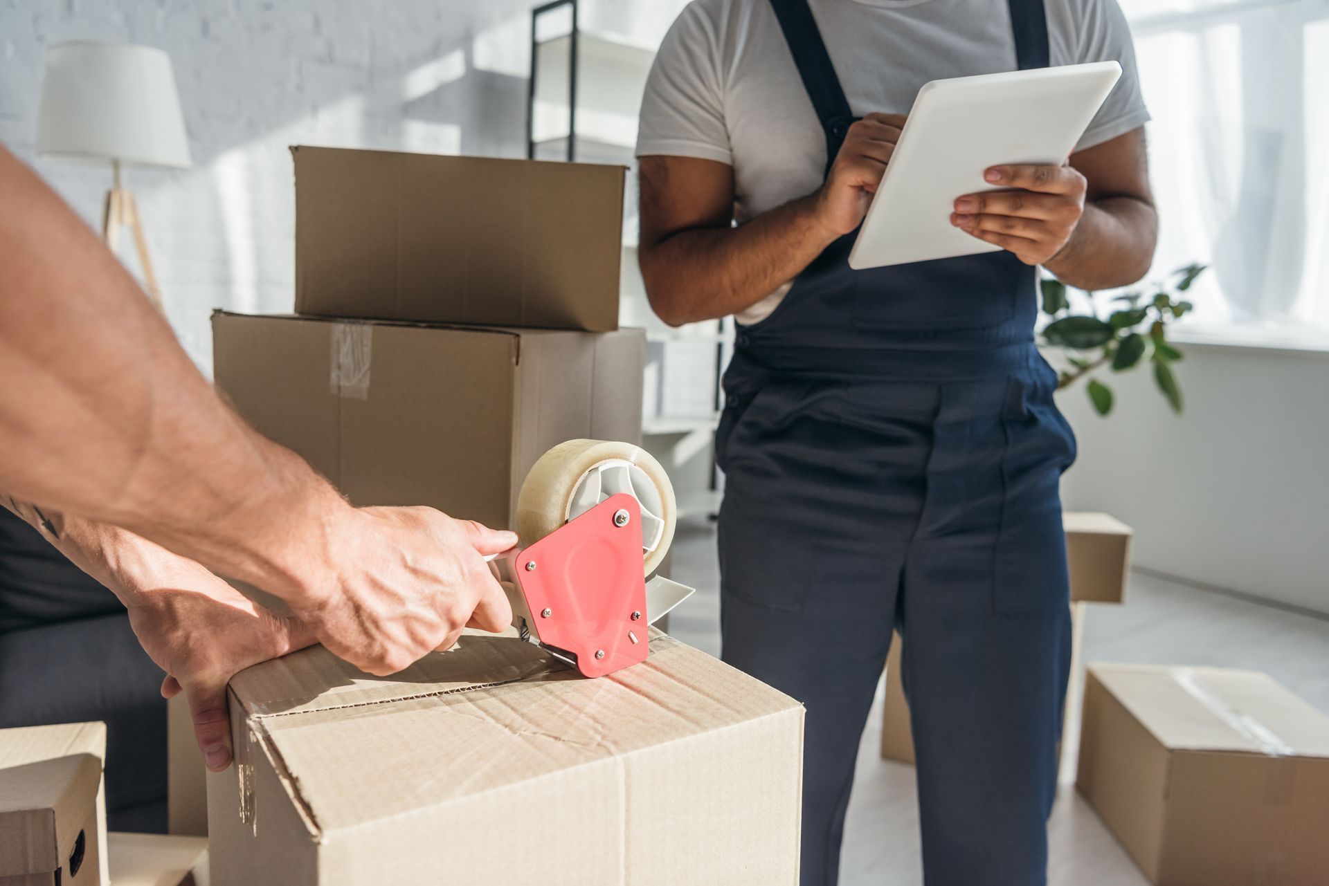 Movers packing boxes, one taping and the other using a tablet in a bright room.