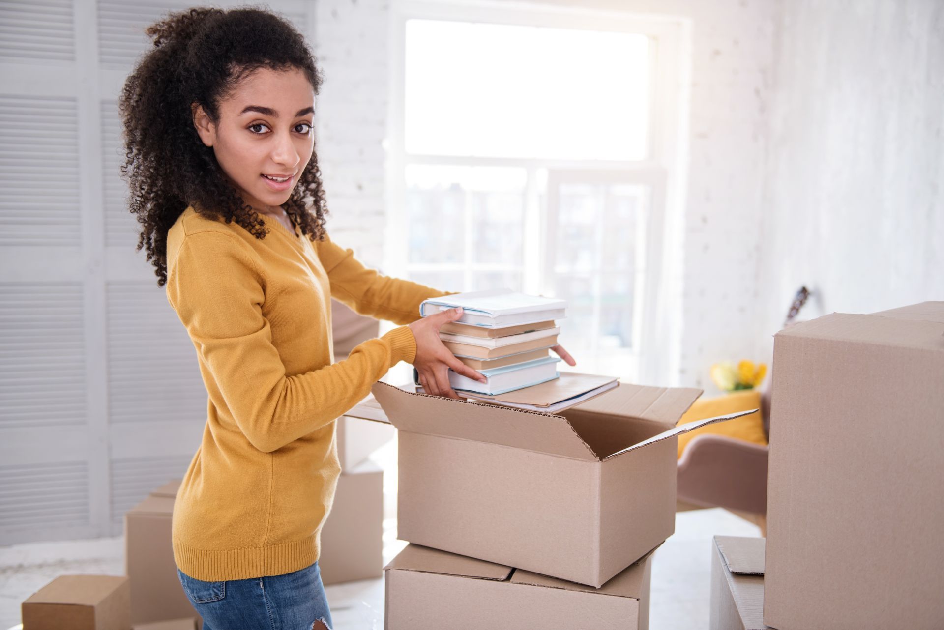 Woman packing books into a moving box in a bright room.