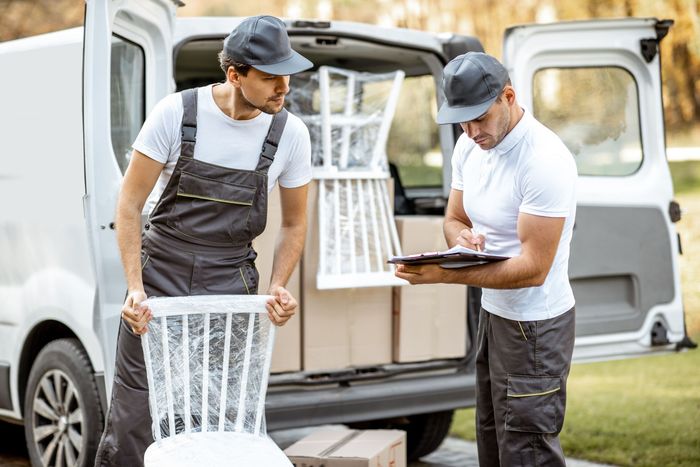 Two movers loading a white van, one wraps a chair, the other reviews a clipboard outdoors.